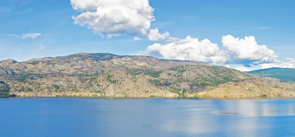 A view of the the mountains across Okanagan Lake under a sunny sky.